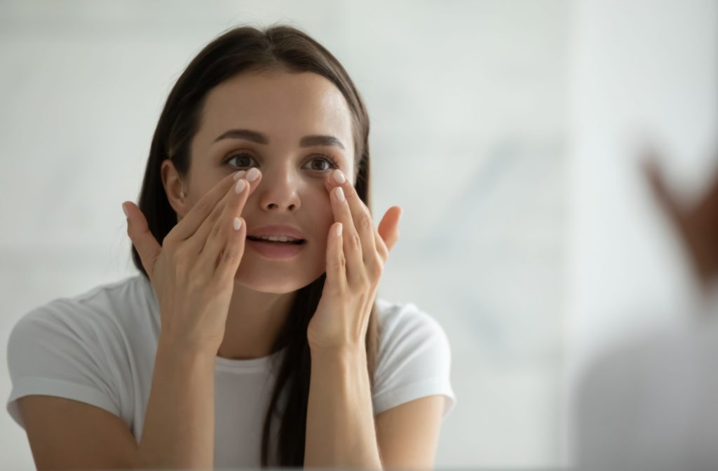 A young adult looks in the mirror as they massage under their eyes with manicured hands to relieve an eye twitch.