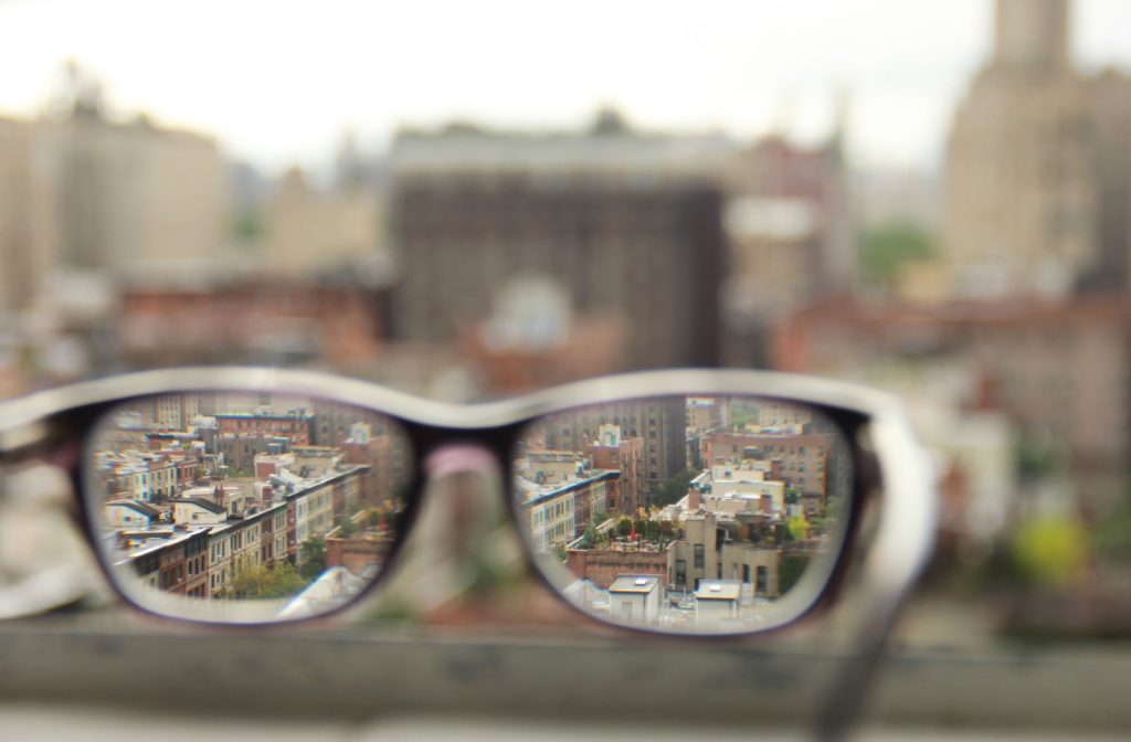 A pair of glasses sitting on a ledge with a view overlooking a blurry city landscape but clear when looking through the glasses lenses