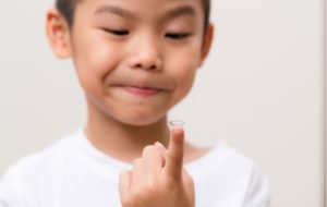 A boy with a white t-shirt holding up a contact lens for his myopia on his fingertip as he looks down at it smiling
