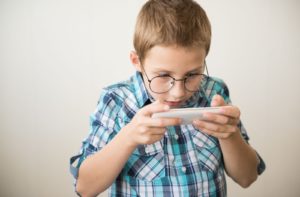 A young boy in glasses is playing a game on a mobile phone with his eyes very close to the screen.