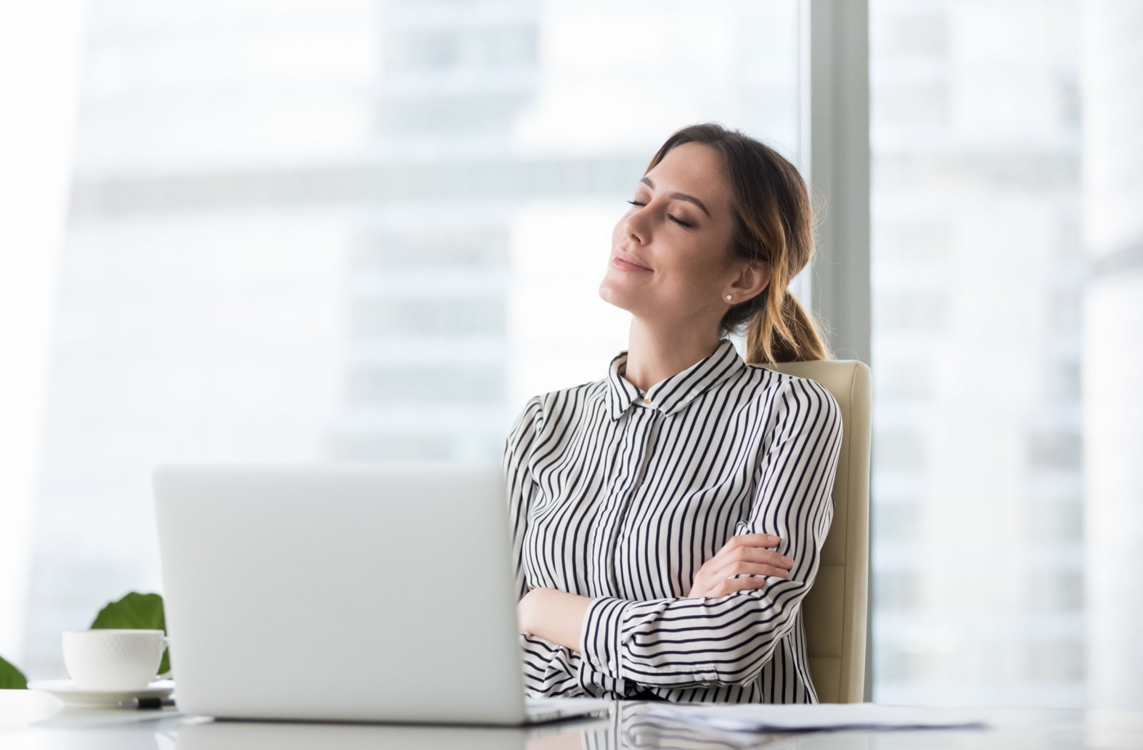 A young woman sitting in front of her laptop in an office, crossing her arms and smiling with her eyes closed