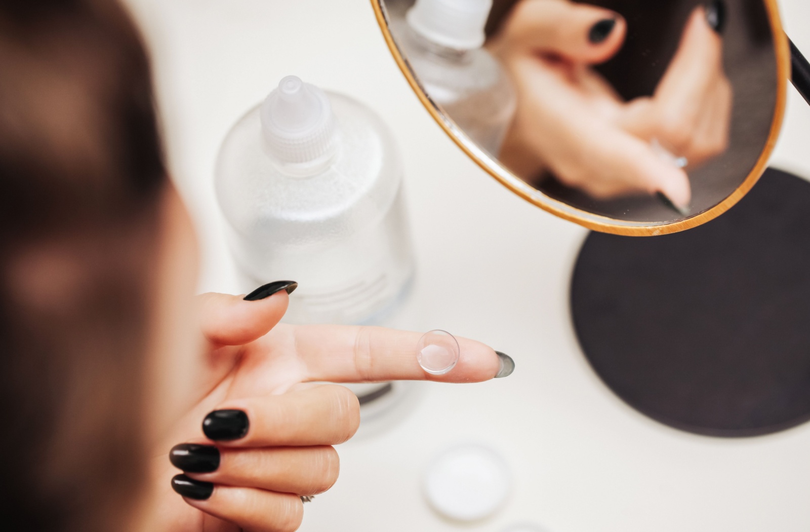A woman with black painted nails balances a contact lens on the index finger of her left hand. A mirror reflects this image.