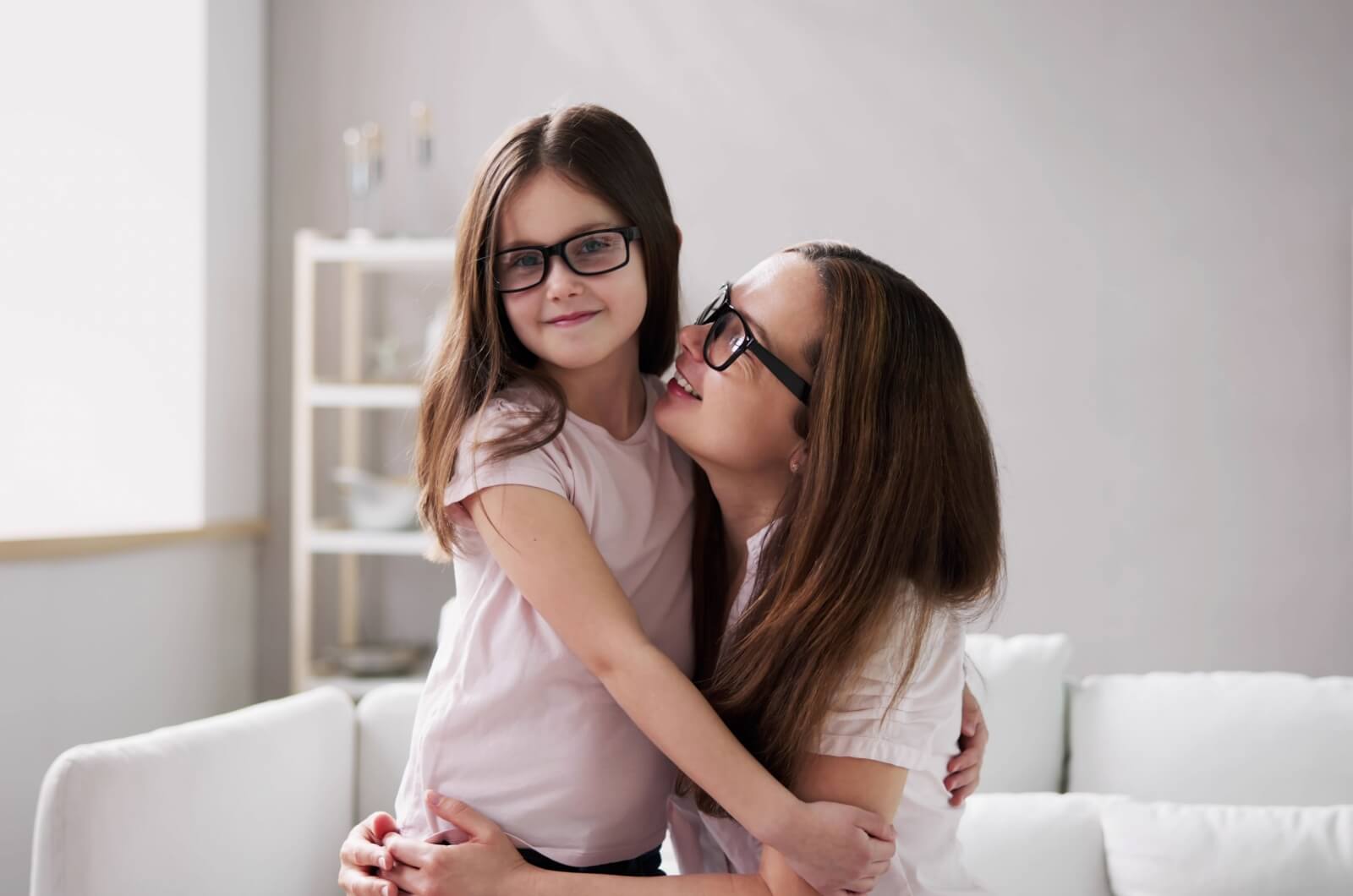 A smiling mother and daughter wearing eyeglasses with arms around each other.