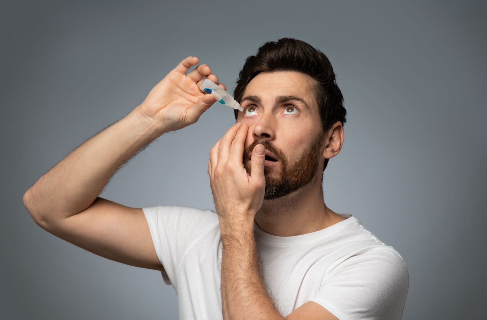 A person applying eye drops against a grey background.