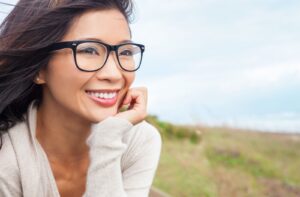 Smiling adult wearing a white cardigan and black-framed glasses sitting outdoors on a grassy hill