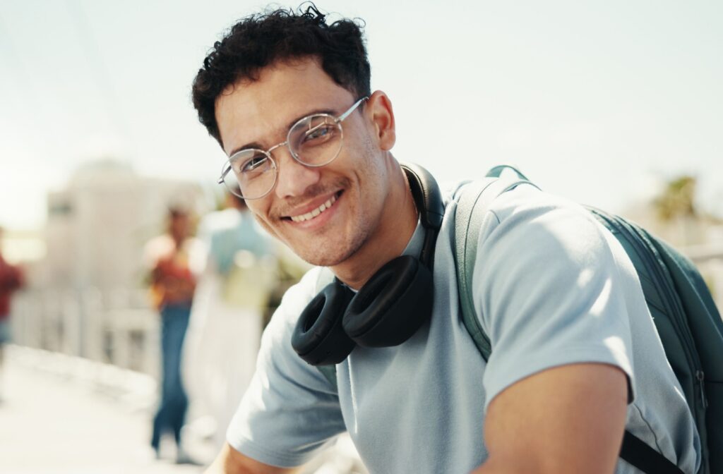 A young adult sitting down wearing glasses, a backpack, and headphones around their neck.