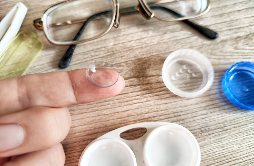 A close up of a contact lens on a finger above a table that has a pair of glasses and a contact lens case on it.