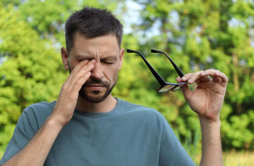 A person rubbing their eyes while holding their glasses in their other hand and standing outside